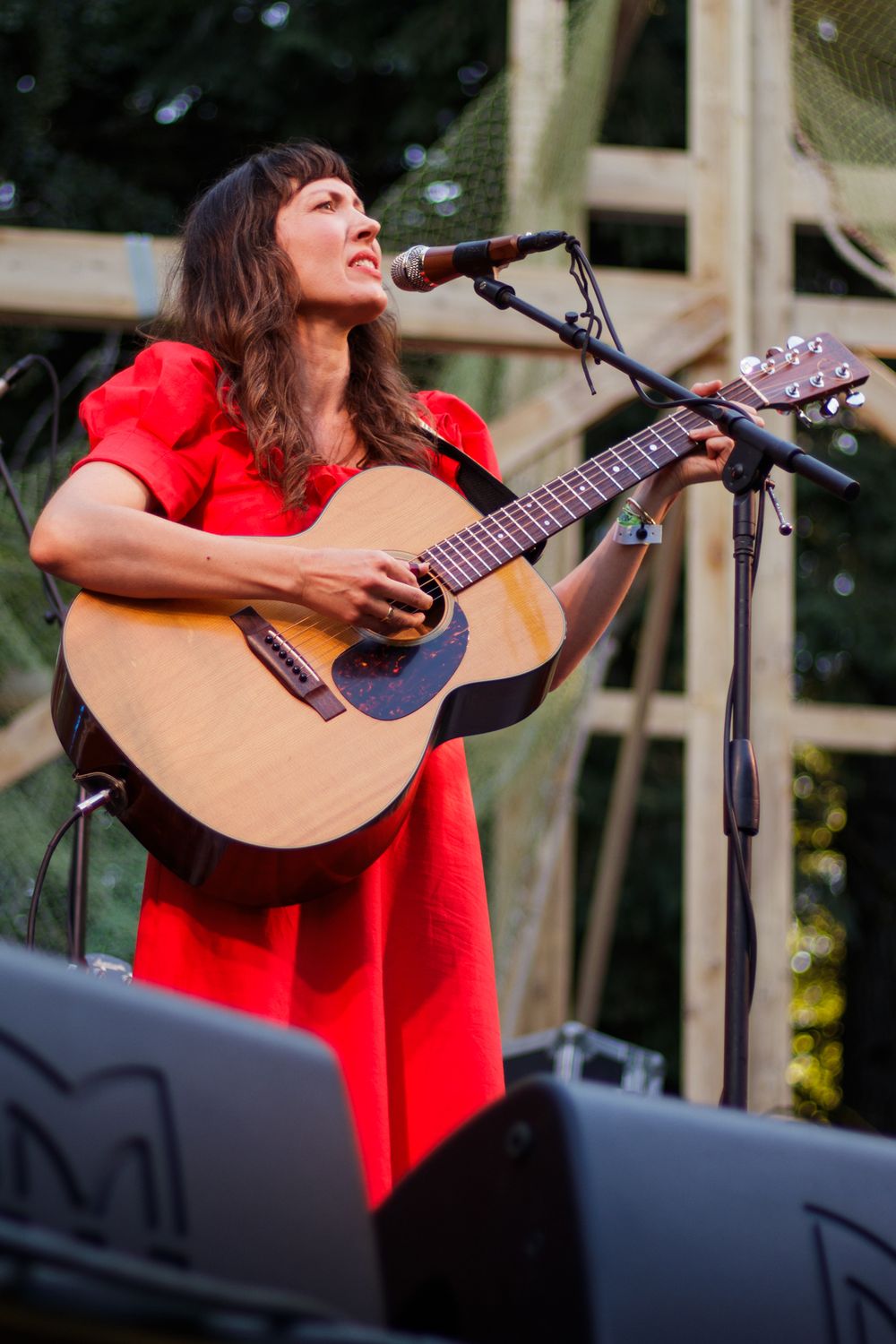 Woman in red dress playing acoustic guitar and singing on outdoor stage.