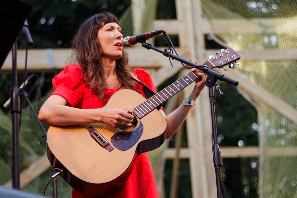 Woman in red dress playing acoustic guitar and singing into microphone on outdoor stage.