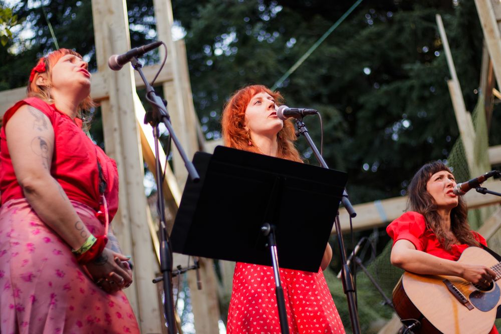 Three women in red perform on outdoor stage, two singing into microphones while one plays acoustic guitar.