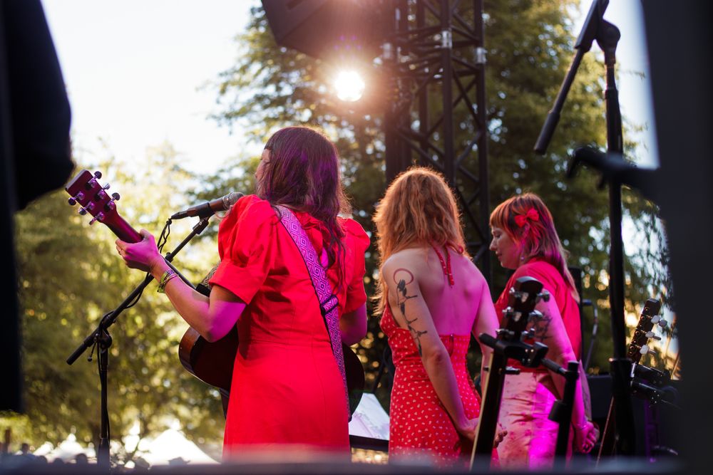 Three female musicians in red dresses perform on outdoor stage, viewed from behind with sunlight filtering through trees.