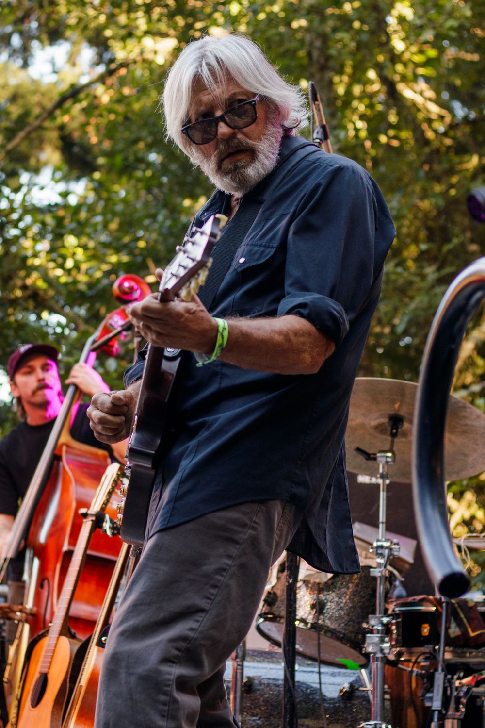 Elderly guitarist with white hair and beard performs outdoors at a music festival, with a bassist visible behind him.