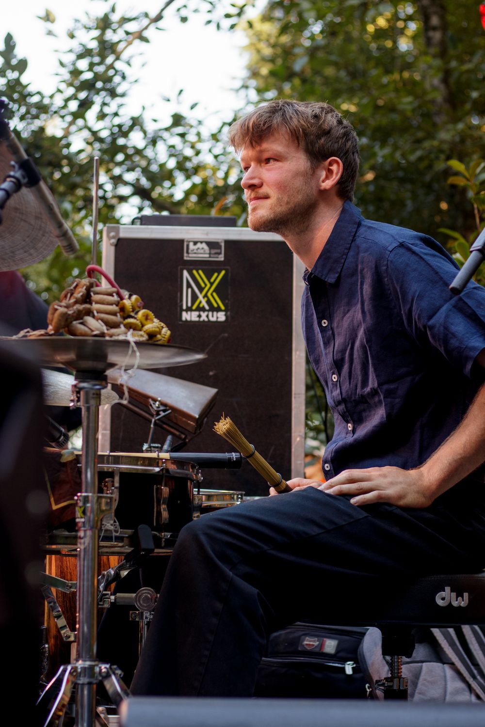 A focused male drummer plays at an outdoor concert with Nexus amplifier equipment visible behind him.