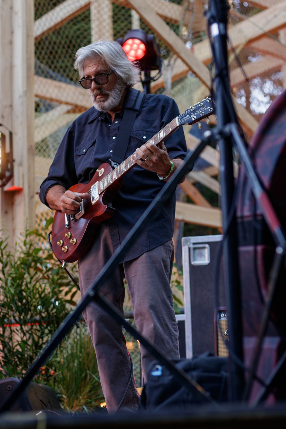 Elderly man with white hair and beard playing a red electric guitar on an outdoor stage.