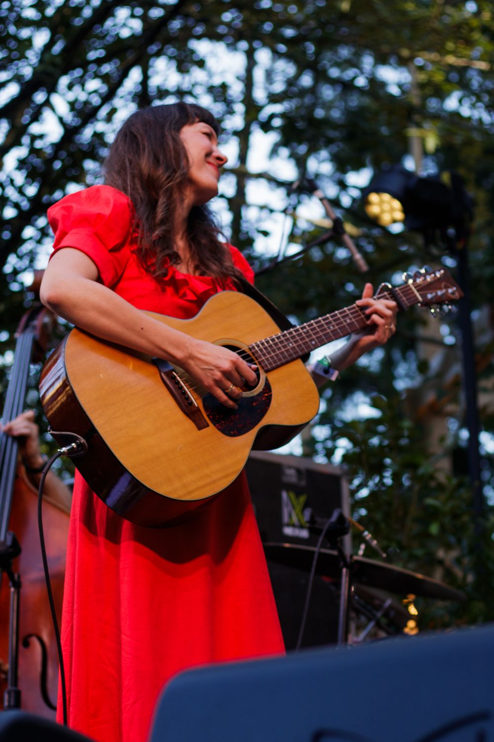 Young woman in a red dress smiling while playing an acoustic guitar on an outdoor stage.