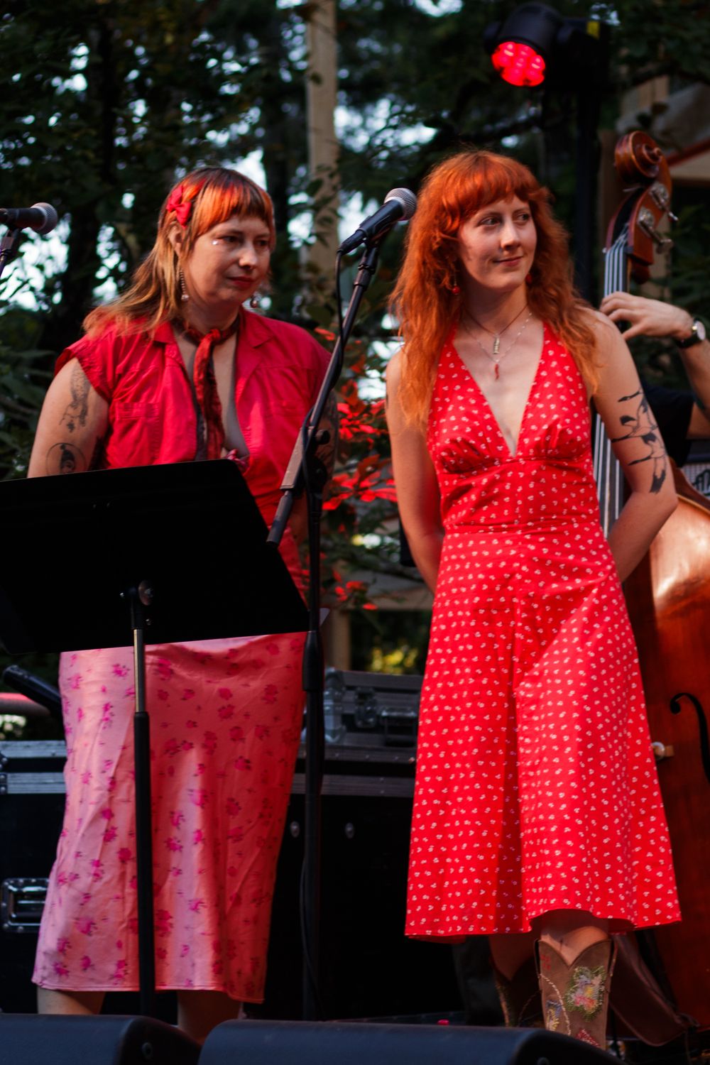 Two women in red outfits perform on an outdoor stage, one in a polka dot dress, the other in a red top and pink skirt.