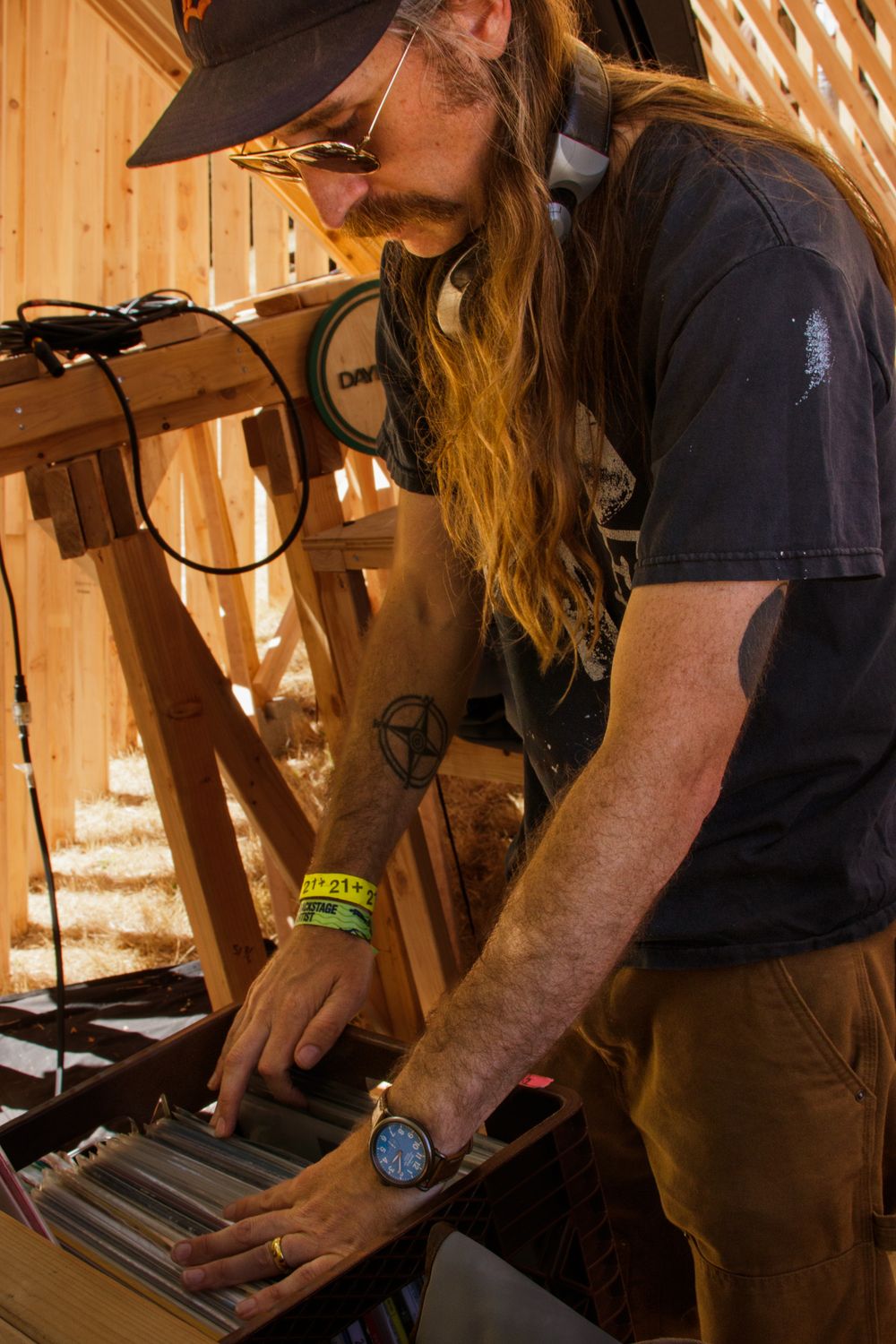 Long-haired DJ browsing vinyl record collection at outdoor wooden DJ booth in sunlight.