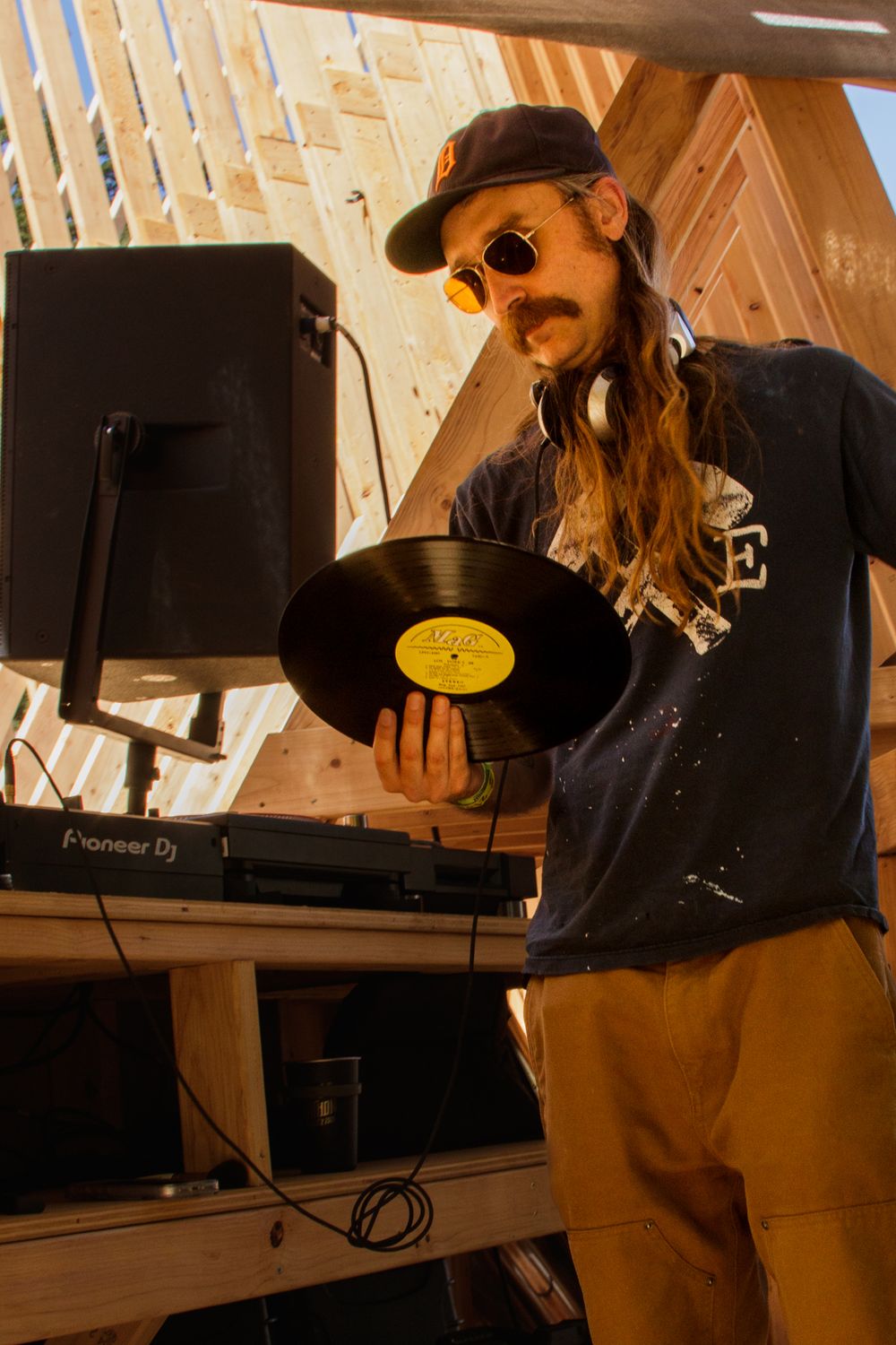 Long-haired DJ holding vinyl record at Pioneer DJ turntable setup in rustic wooden venue.