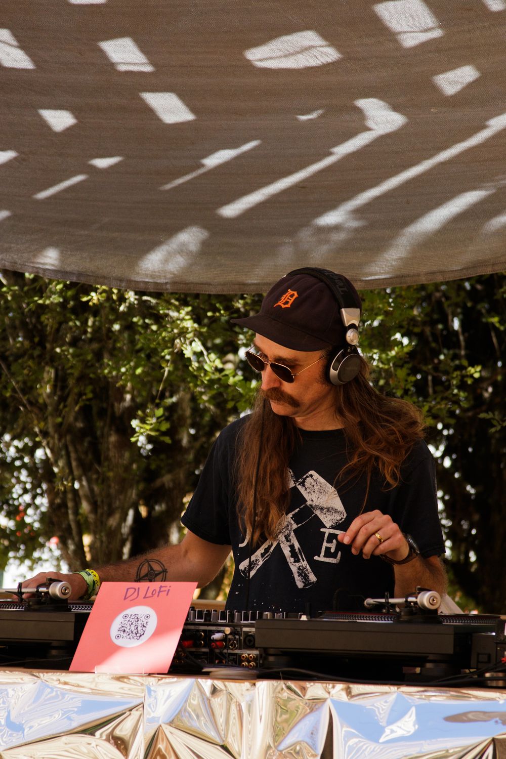A DJ with long hair, sunglasses, and headphones mixing records outdoors under a shaded canopy at a music festival.