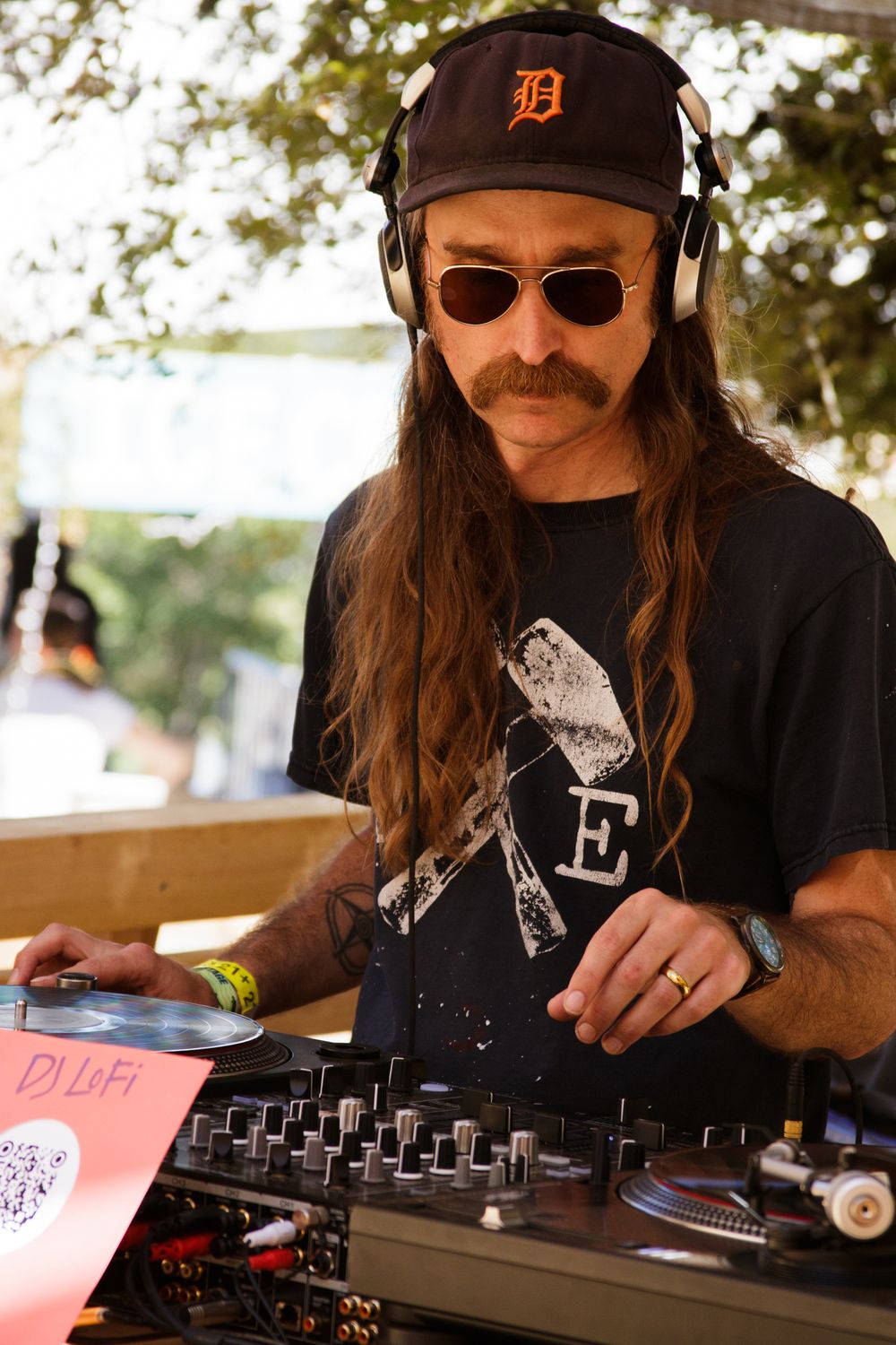 DJ LoFi mixing records outdoors, sporting Detroit Tigers cap, headphones, sunglasses, and mustache.