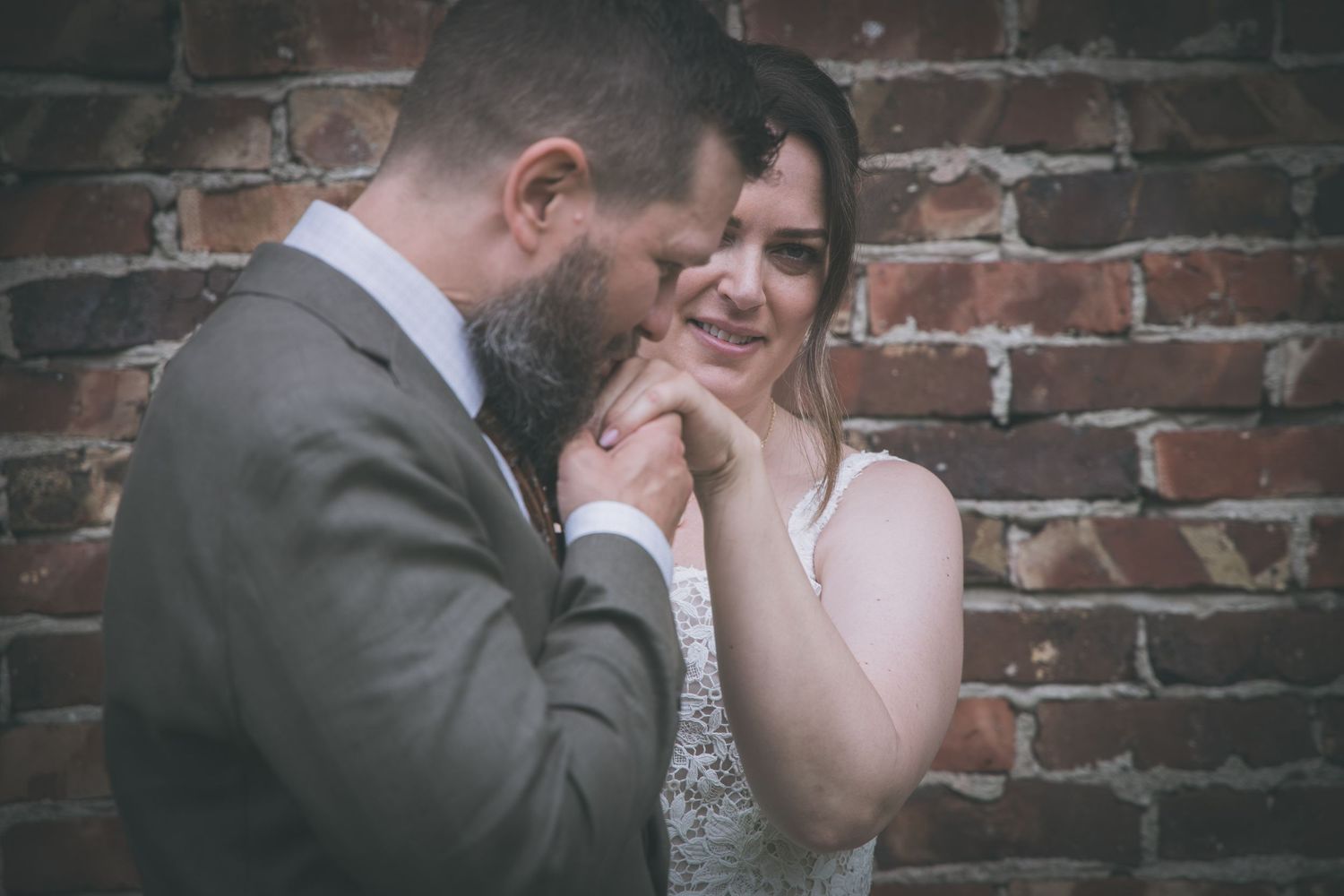 Groom kisses bride's hand as she smiles joyfully against a rustic brick wall background.