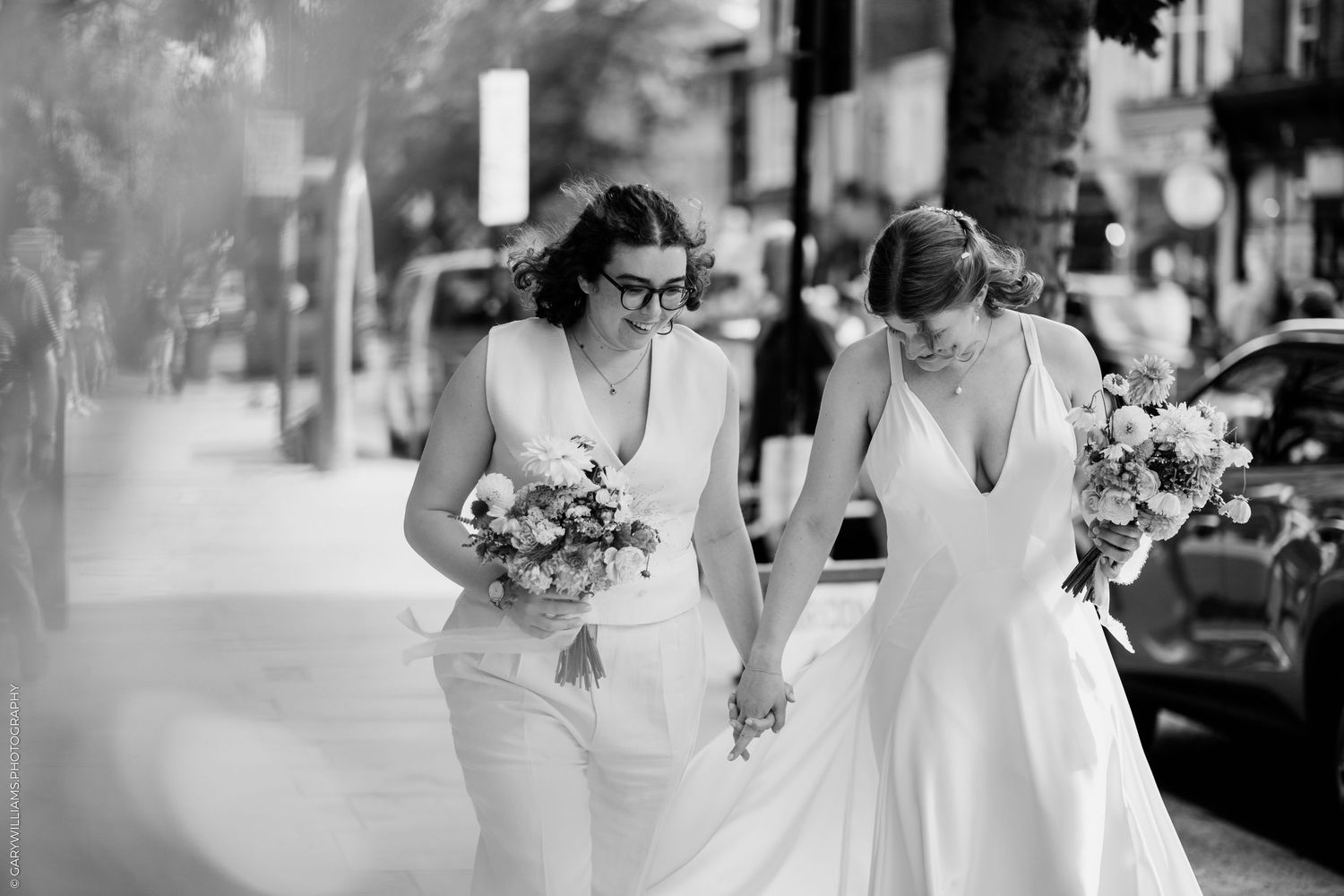 Two brides in white holding bouquets walk hand in hand on a city sidewalk in this black and white wedding photo.