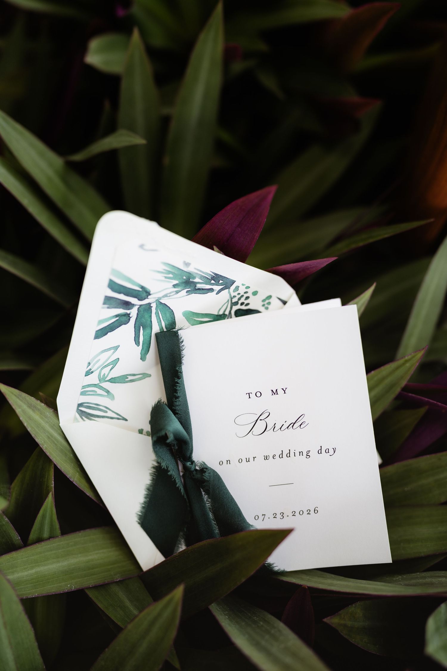 Elegant wedding card addressed to the bride resting on lush tropical green leaves with a botanical envelope liner.