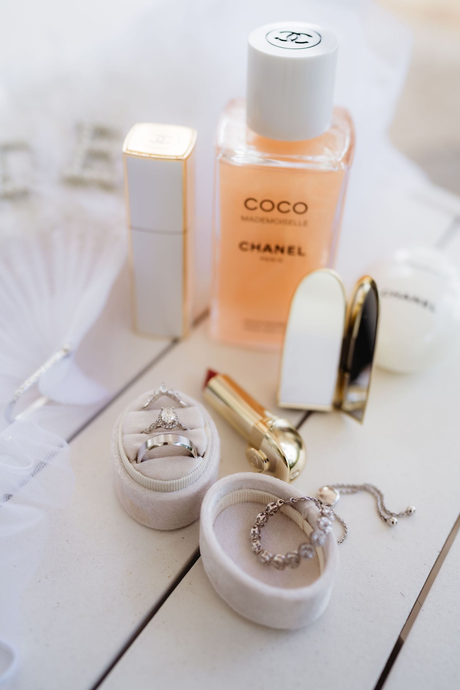 Chanel Coco Mademoiselle perfume with diamond rings in velvet box, gold lipstick, and luxury beauty accessories on white surface.