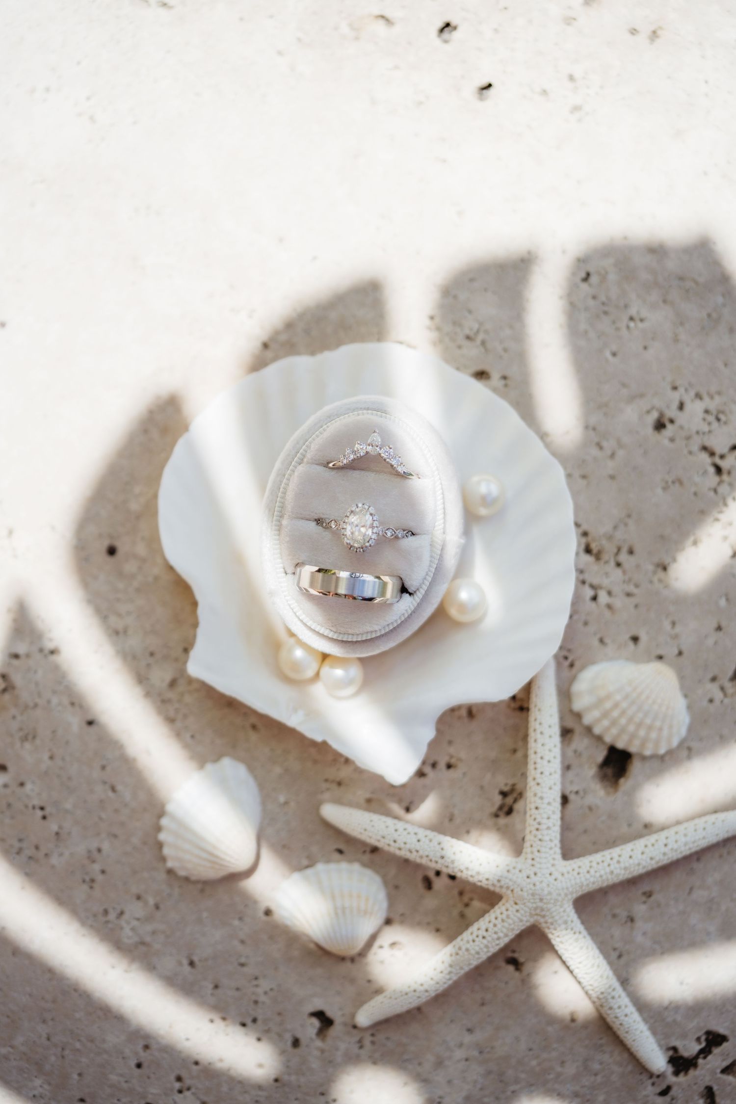 Wedding rings displayed in a velvet box on a seashell surrounded by pearls and starfish on sandy beach.
