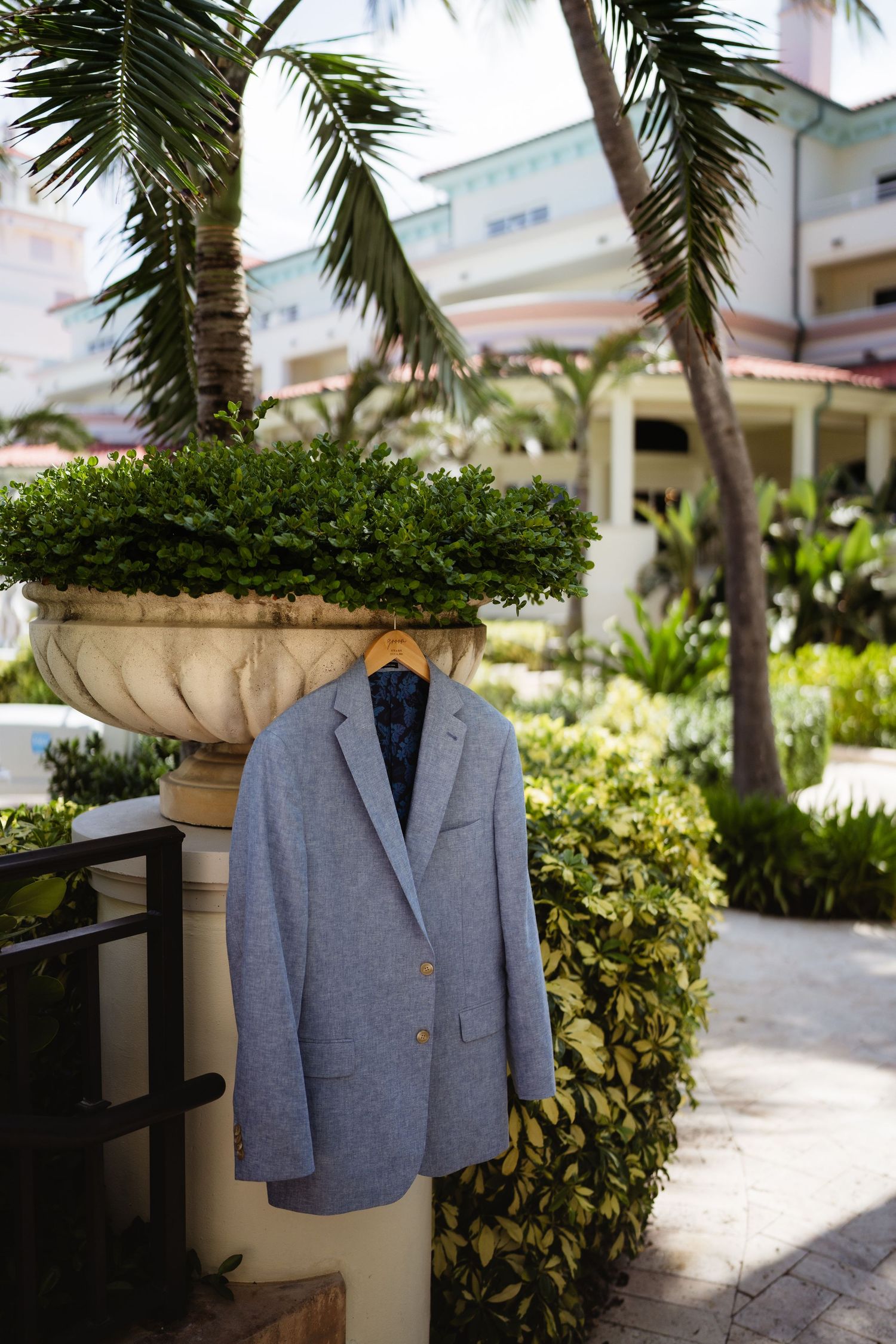 Gray blazer on wooden hanger displayed outdoors near tropical palm trees and lush greenery at a resort.