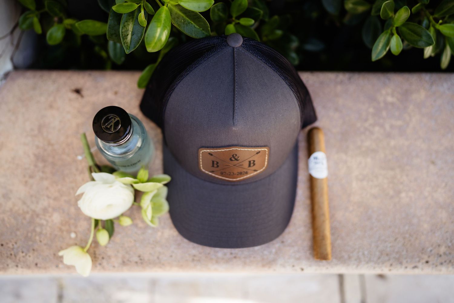 Dark gray trucker hat with leather patch beside a cigar and bottle on stone surface with green foliage backdrop.