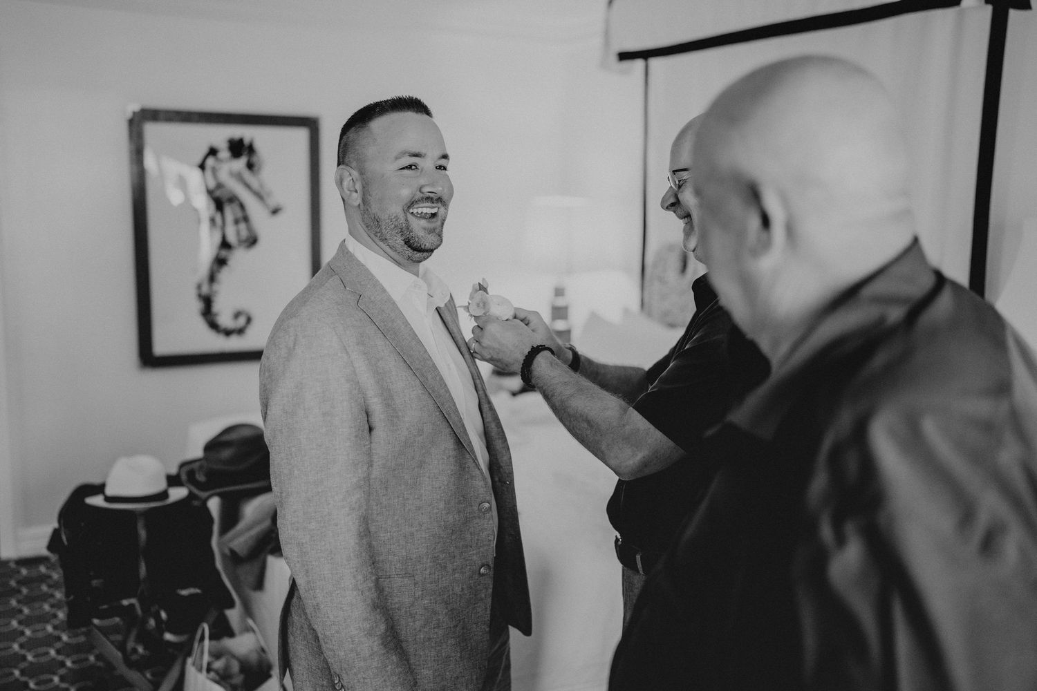 Black and white photo of a groom laughing as a man adjusts his suit jacket before a wedding ceremony.