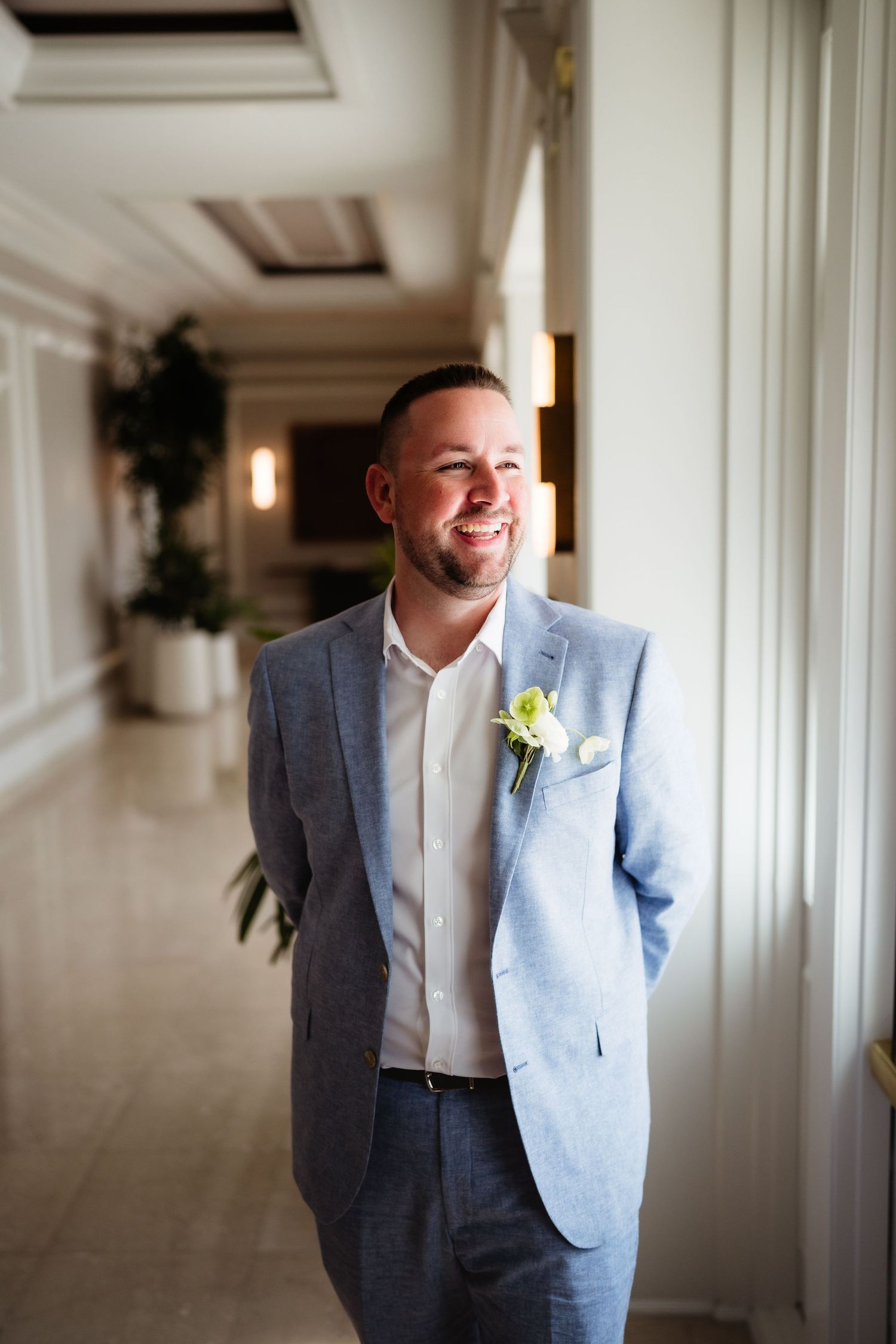 Smiling groom in light blue suit with white boutonniere standing in elegant hotel corridor.
