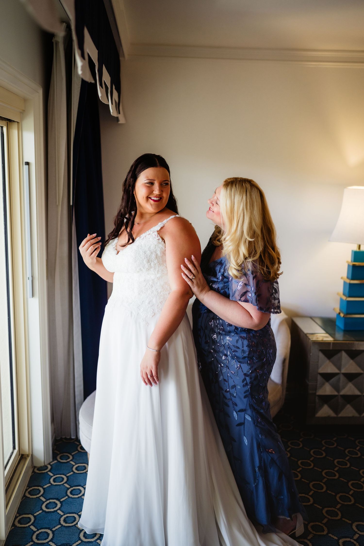 Bride in white lace gown smiles as blonde woman in blue dress helps her prepare in a hotel room.