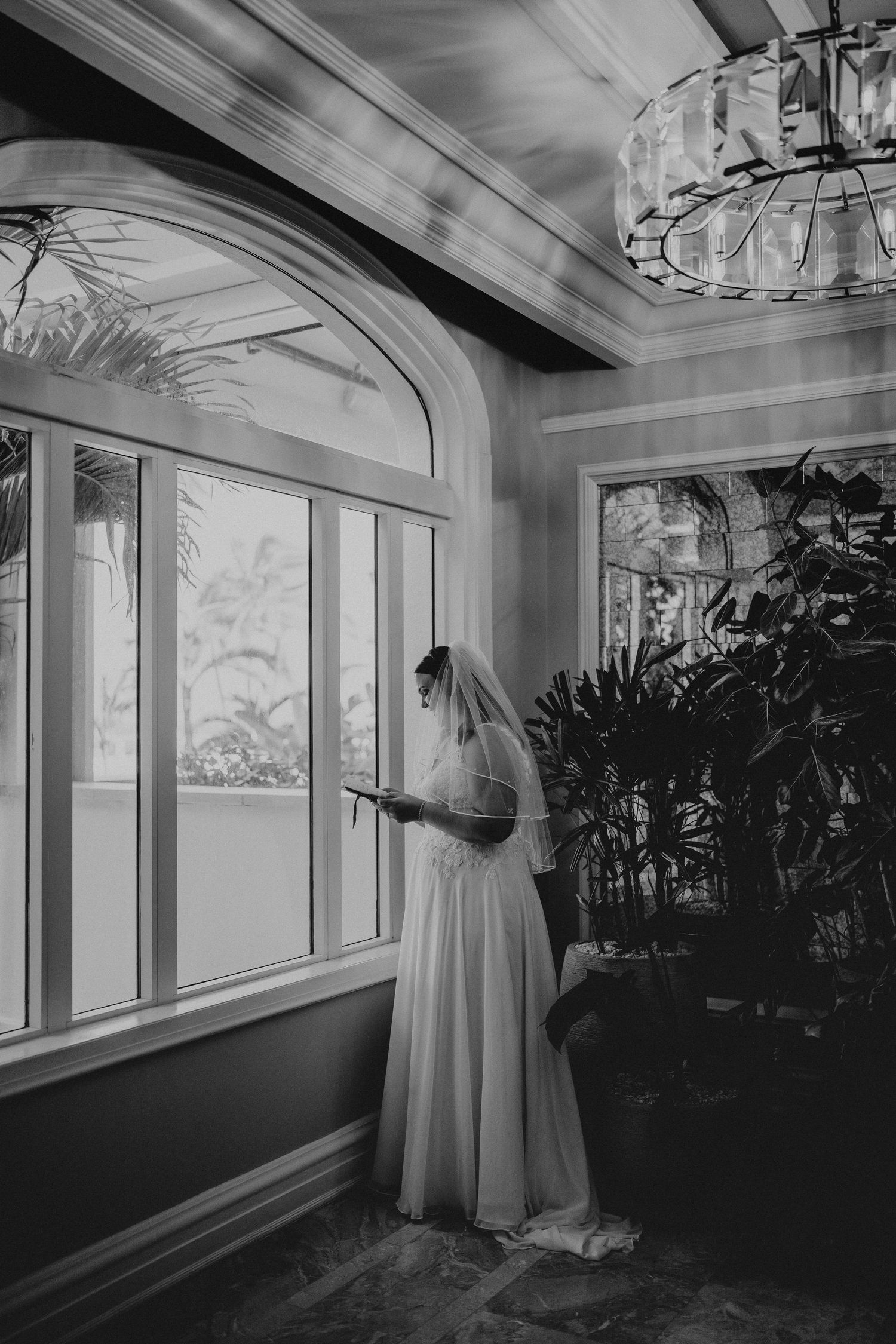 Black and white photo of a bride in a wedding dress and veil reading a note by large arched windows in an elegant venue.