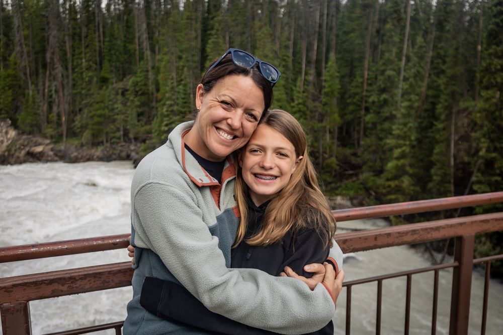 Two people embrace and smile while standing on a wooden bridge in a scenic forest setting with evergreen trees.
