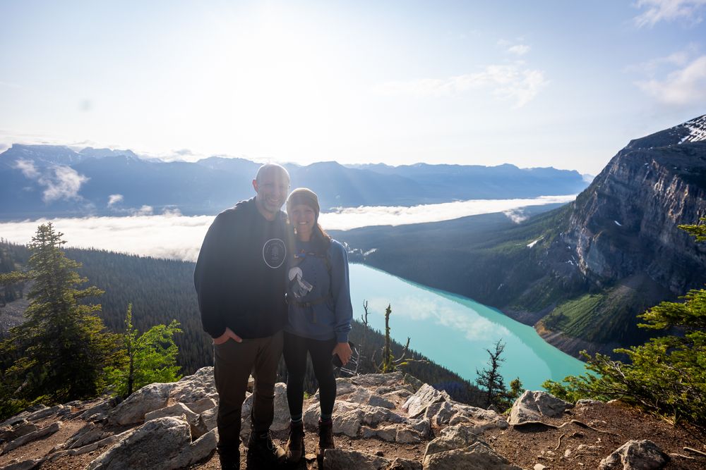 Two hikers stand on a mountain viewpoint overlooking a turquoise alpine lake surrounded by mountains and forest.