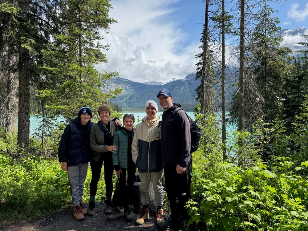 A group of hikers pose on a forest trail overlooking a turquoise mountain lake on a sunny summer day.