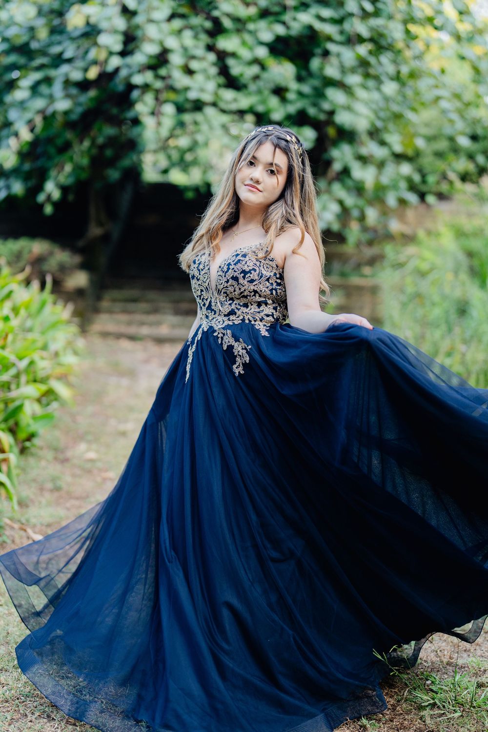 Young woman in elegant navy blue ball gown with gold embroidery poses gracefully outdoors in a lush garden setting.