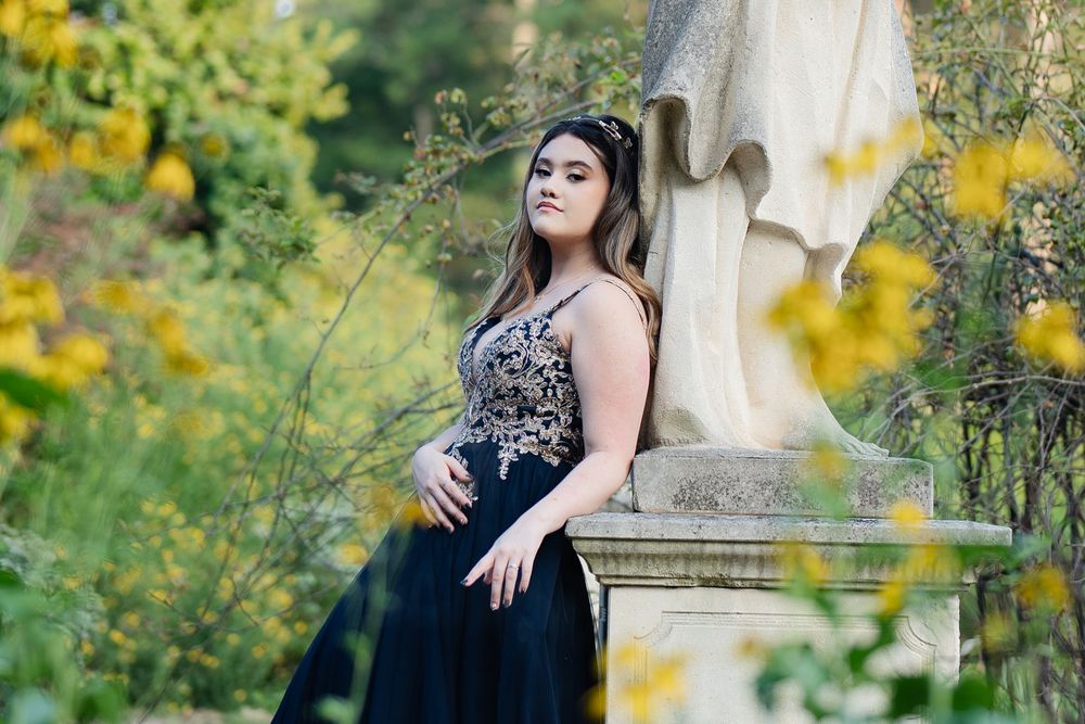 Young woman in elegant black gown poses near stone statue surrounded by lush yellow wildflowers.