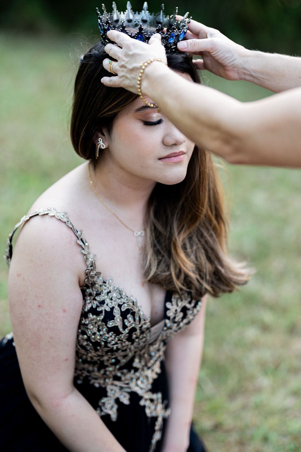 Young woman in black gold gown being crowned with blue tiara outdoors during pageant ceremony.