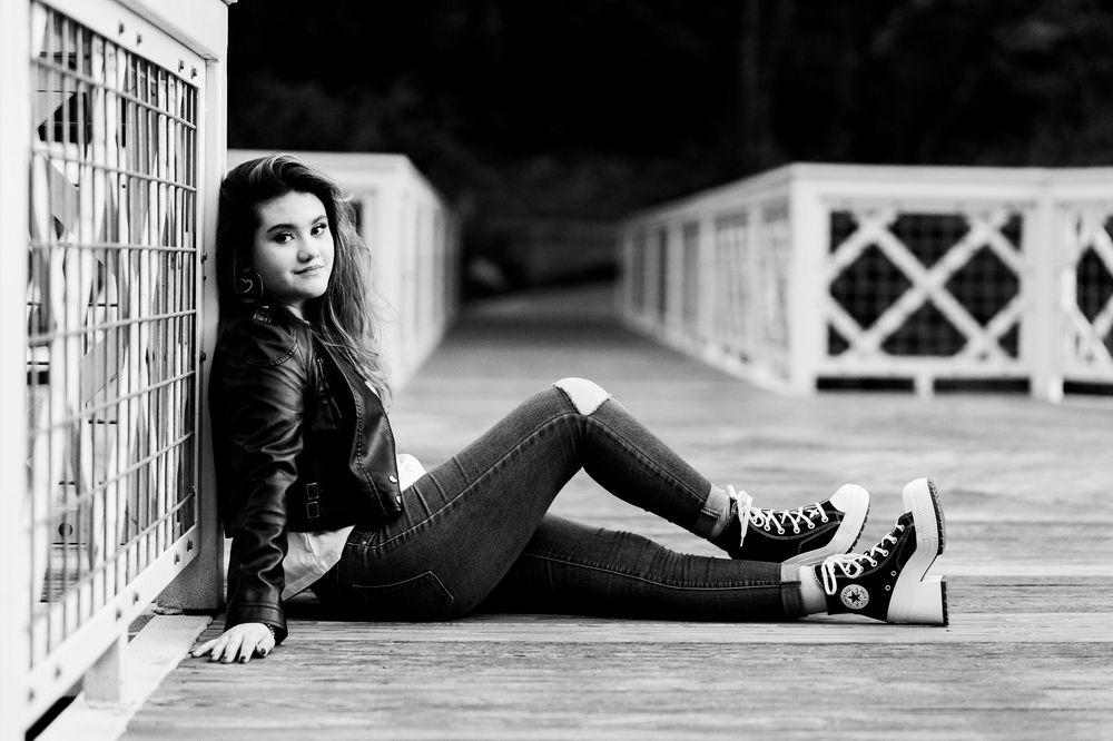 Young woman in black and white photo sitting on bridge in leather jacket and Converse sneakers.