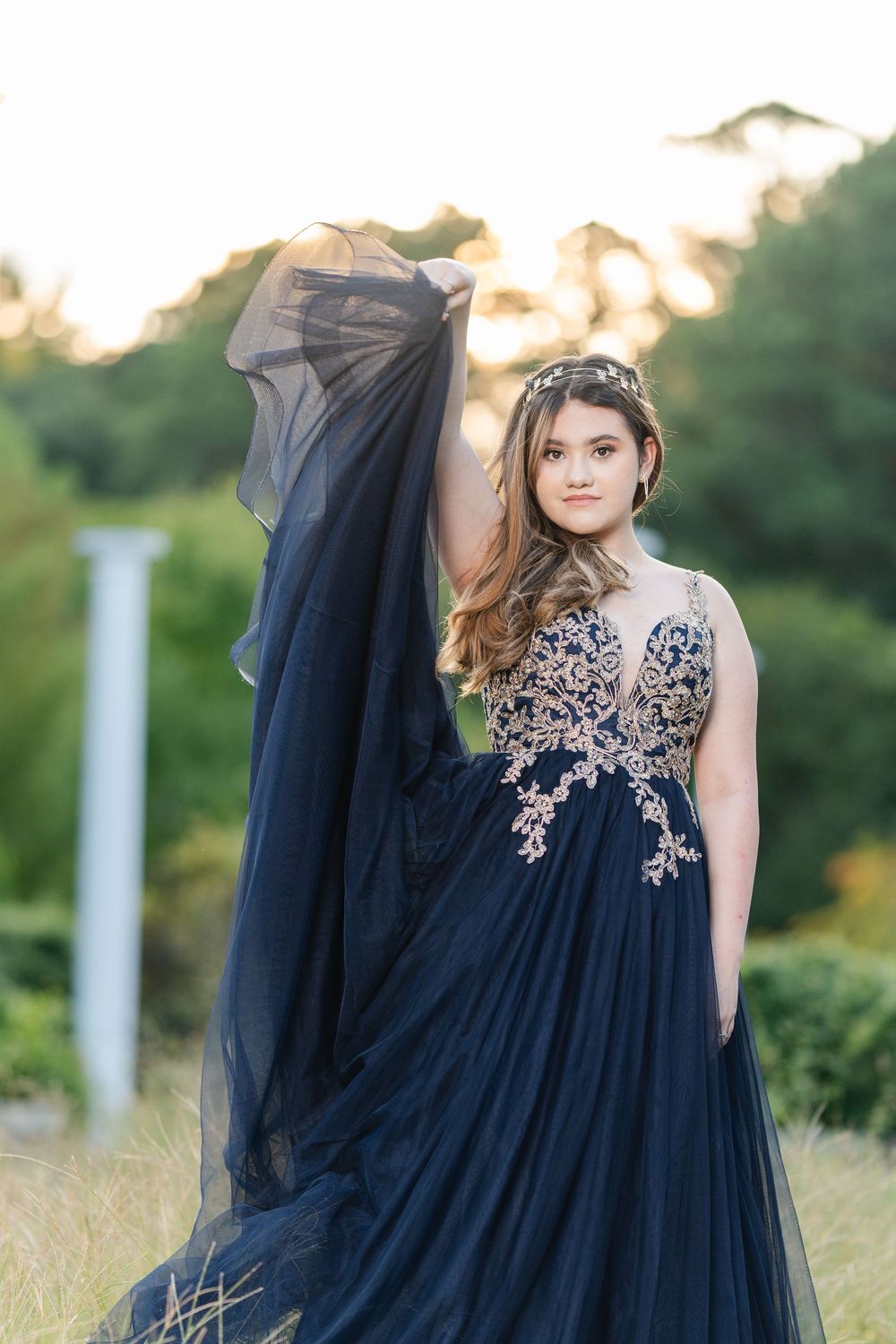 Young woman in navy blue ball gown with gold lace bodice poses outdoors at golden hour, holding flowing tulle skirt.