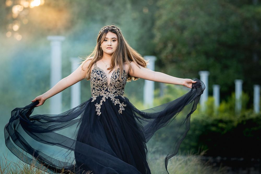 Young woman in elegant black ball gown with gold embroidery poses gracefully outdoors in soft natural light.