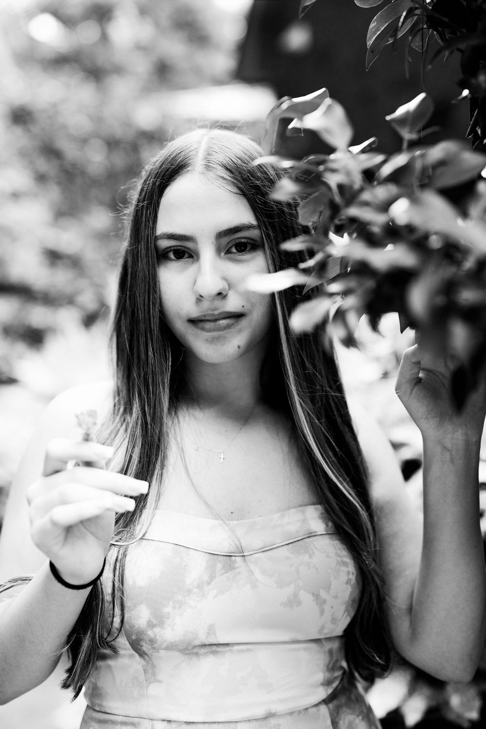 Black and white portrait of a young woman with long hair standing near leafy branches outdoors.