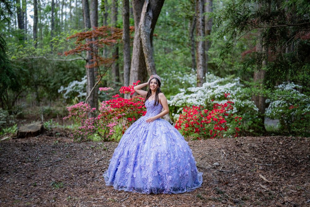 Young woman in lavender ball gown poses among colorful azalea blooms in a lush forest garden setting.