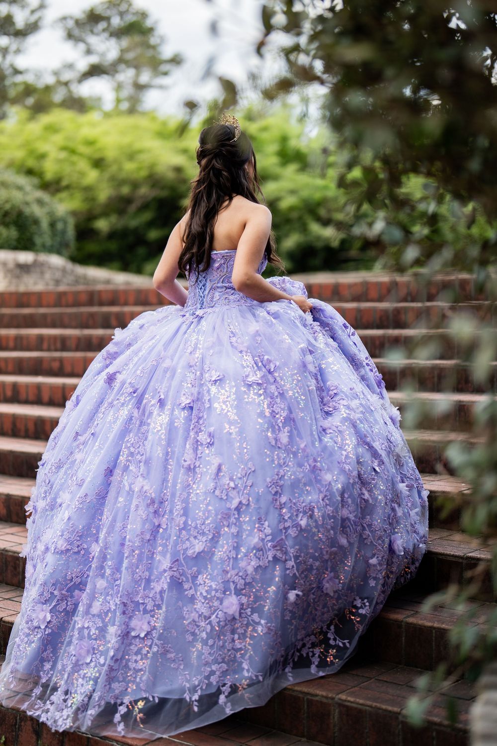 Young woman in lavender floral ball gown quinceañera dress ascending brick stairs in a lush garden setting.