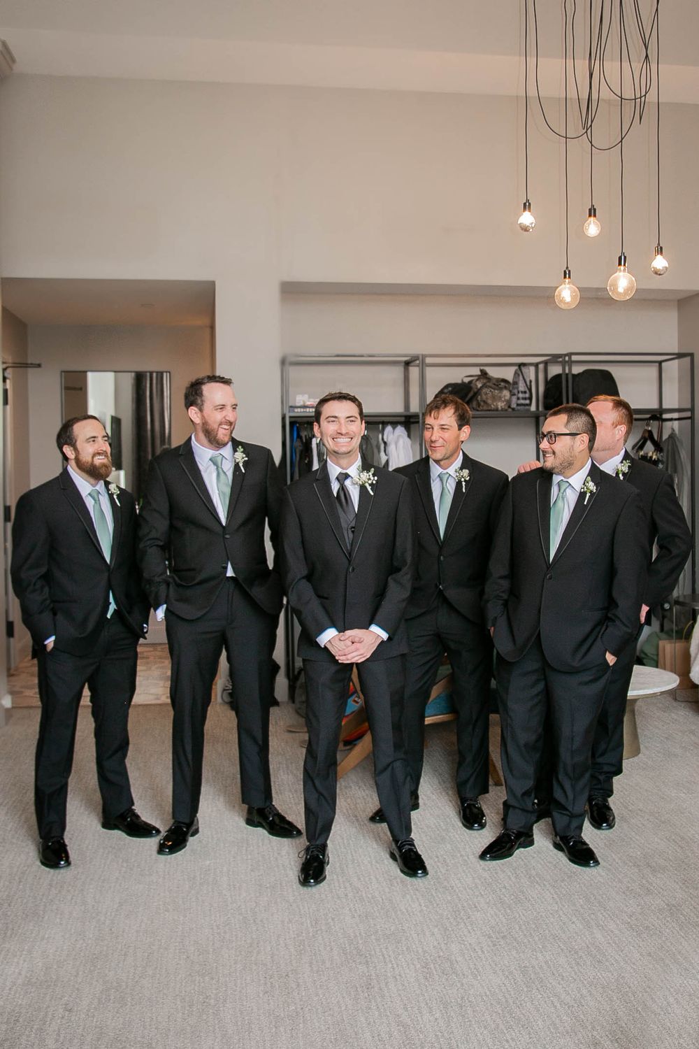 Groomsmen in matching black suits and mint green ties stand together in modern room with hanging pendant lights.