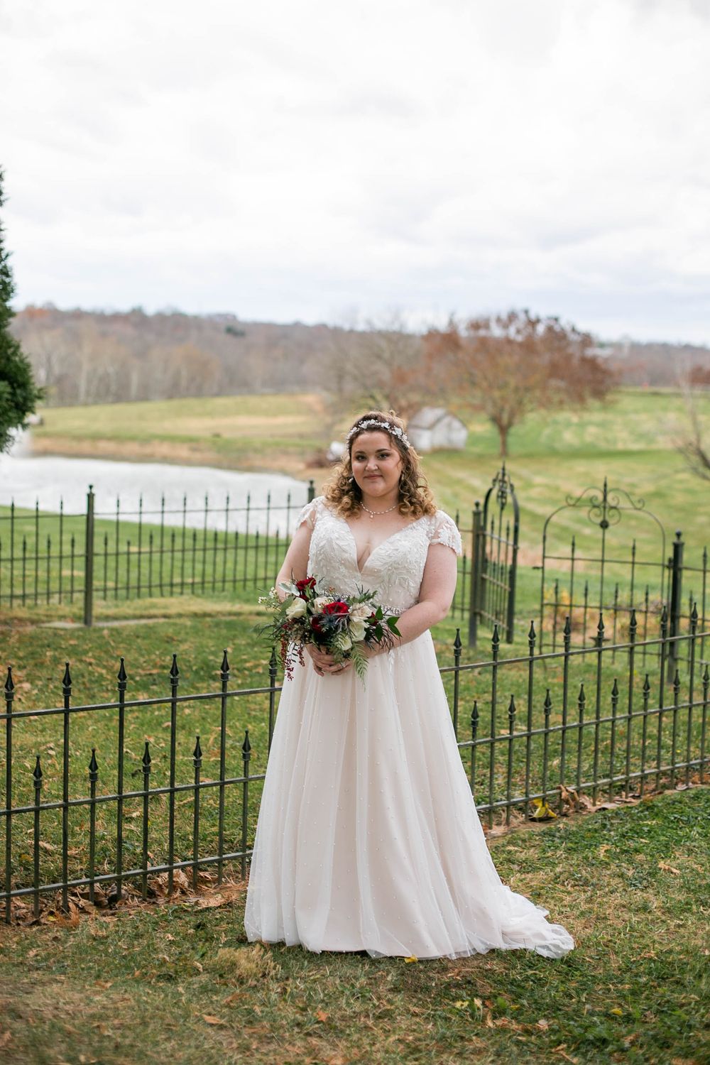 A bride in a flowing white gown stands by an ornate iron fence overlooking a scenic countryside landscape with rolling hills.