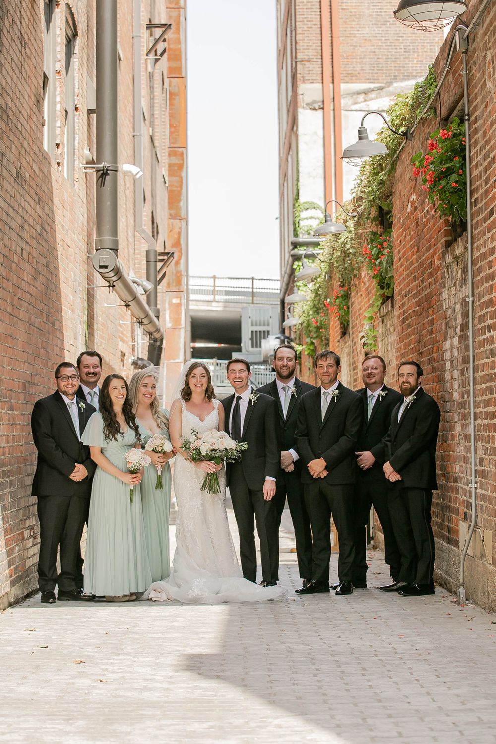 A bridal party poses together in an alleyway between brick buildings with greenery growing on the walls.