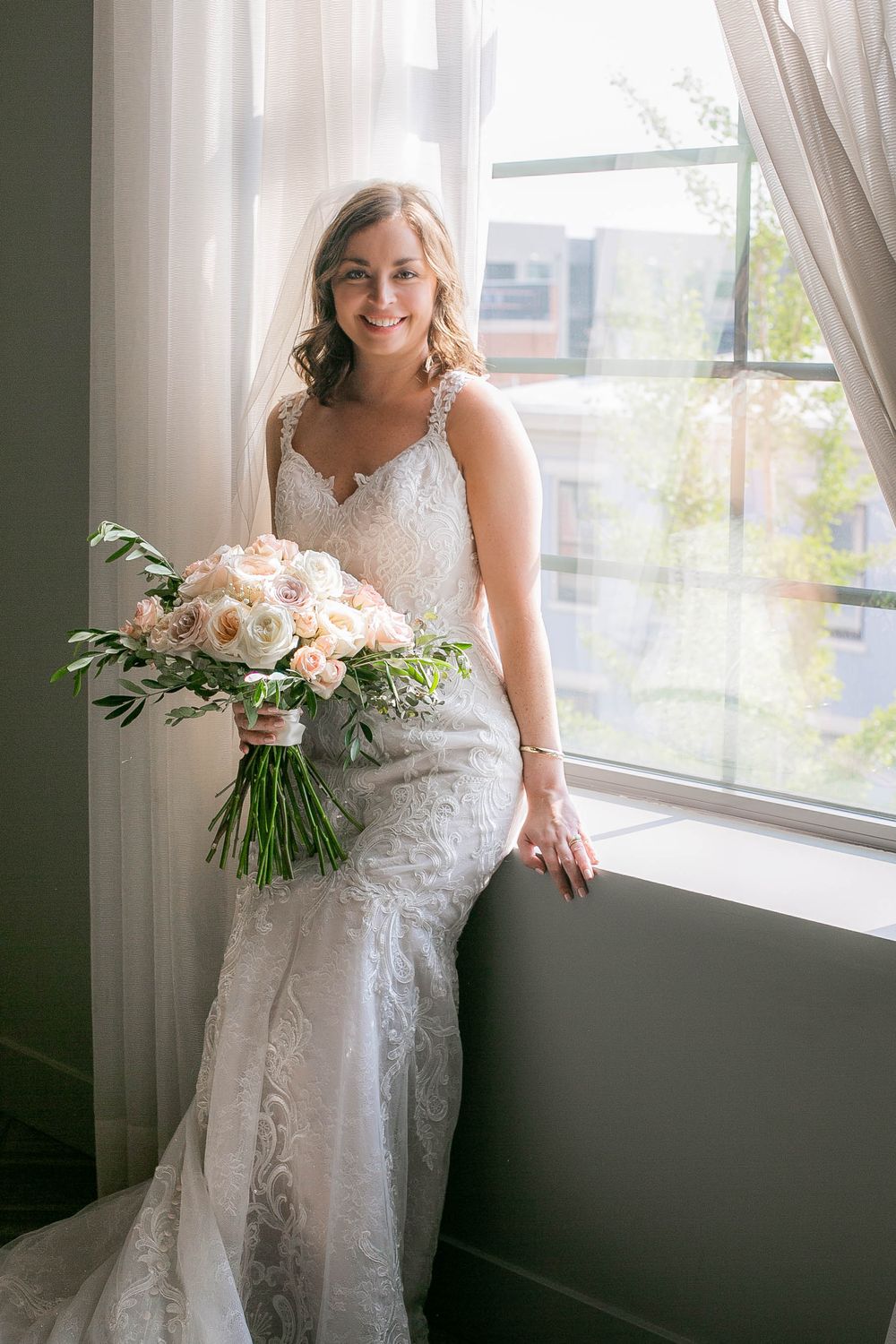 Elegant lace wedding dress with delicate straps accentuated by soft natural light and blush pink bouquet by window.