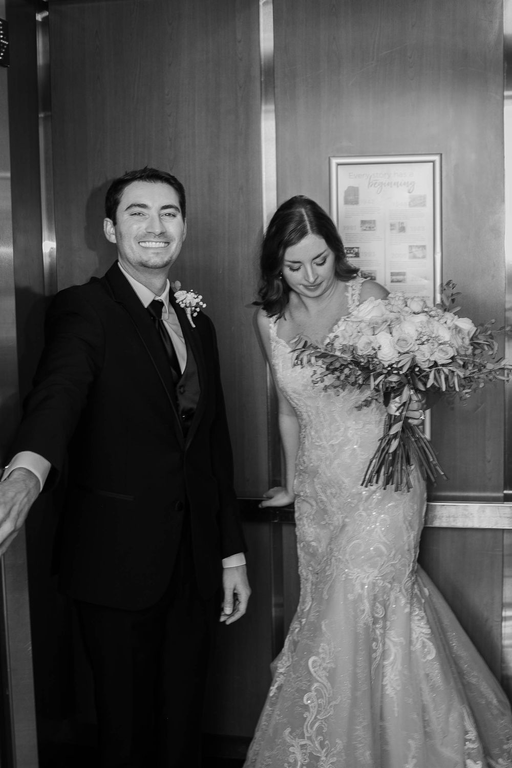 A black and white wedding photo showing a couple in an elevator, the bride holding a large floral bouquet.