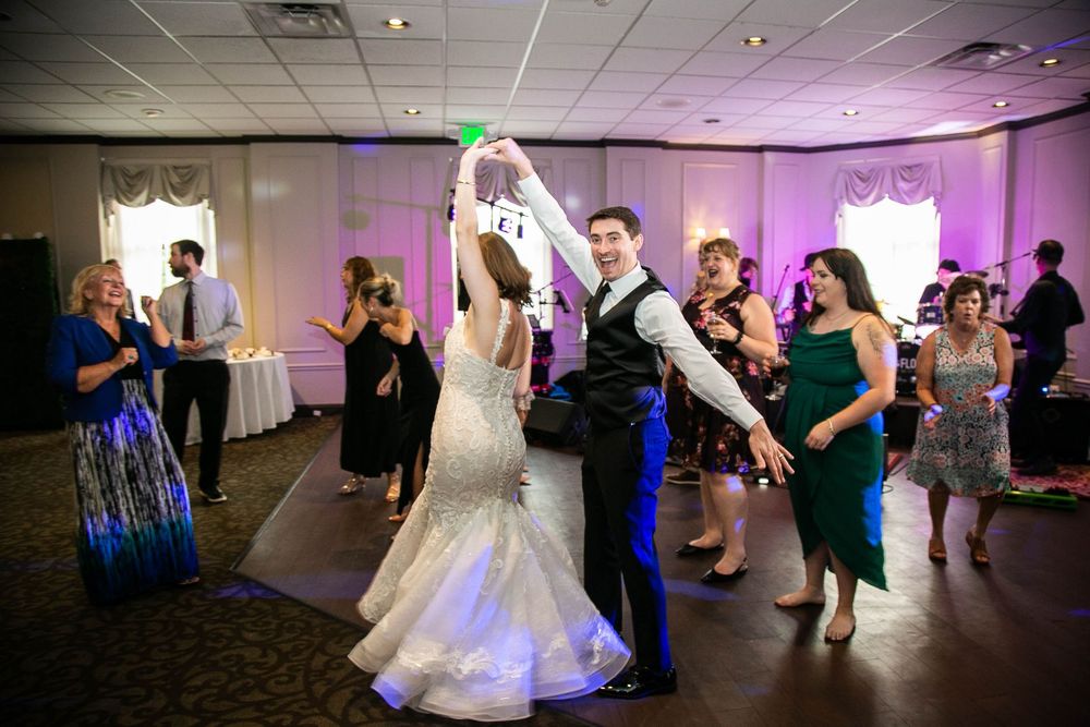 Wedding guests dance and celebrate during a reception in a ballroom with purple lighting and white decor.