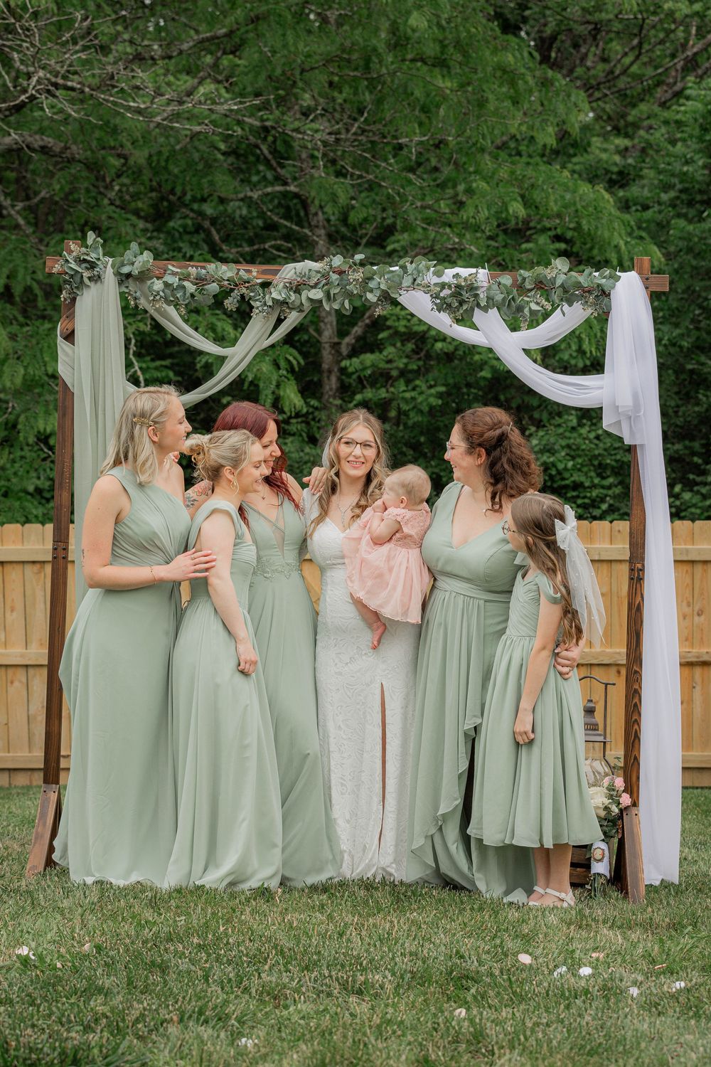 Bridesmaids in sage green dresses and a bride in white share candid laughing moments together on a grassy lawn.