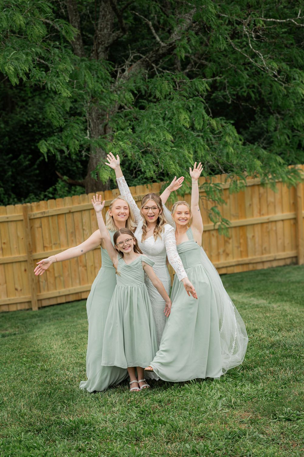 Group in sage green dresses poses playfully on grass near wooden fence during outdoor photo session.