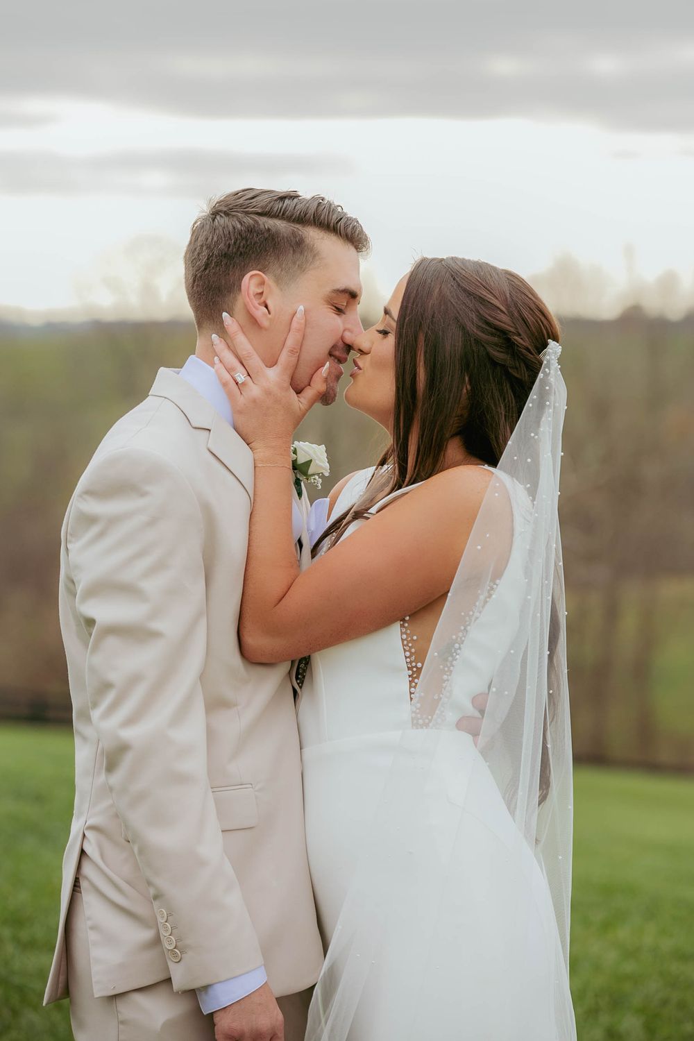 A bride and groom share a romantic kiss on their wedding day on a grassy hill under cloudy skies.