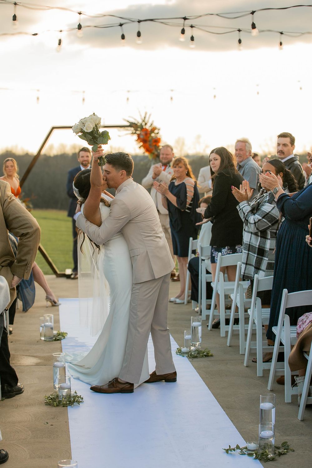 A couple shares their first kiss during a sunset outdoor wedding ceremony while guests applaud on a white aisle runner.