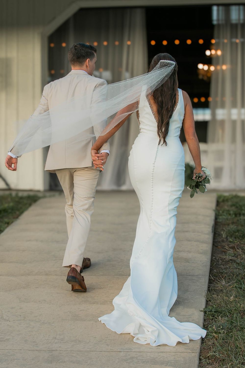 A couple walks away holding hands on their wedding day, bride in white fitted gown with veil and groom in light beige suit.