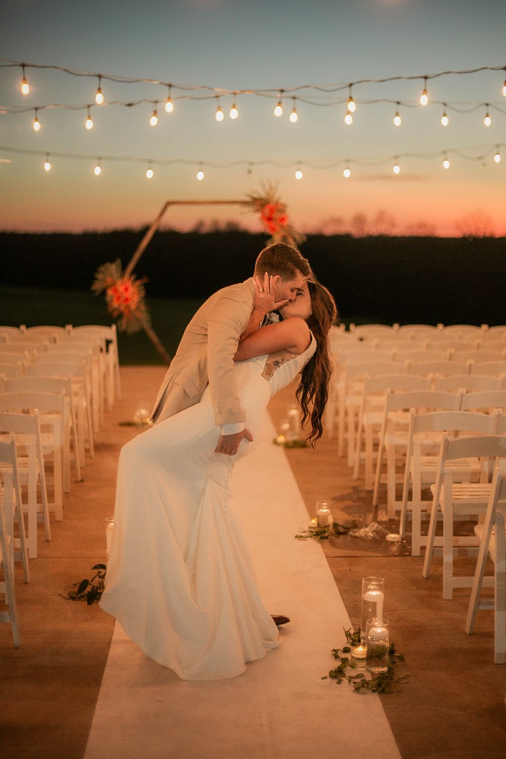 A wedding ceremony setup at sunset with string lights overhead and white chairs along a decorated aisle.