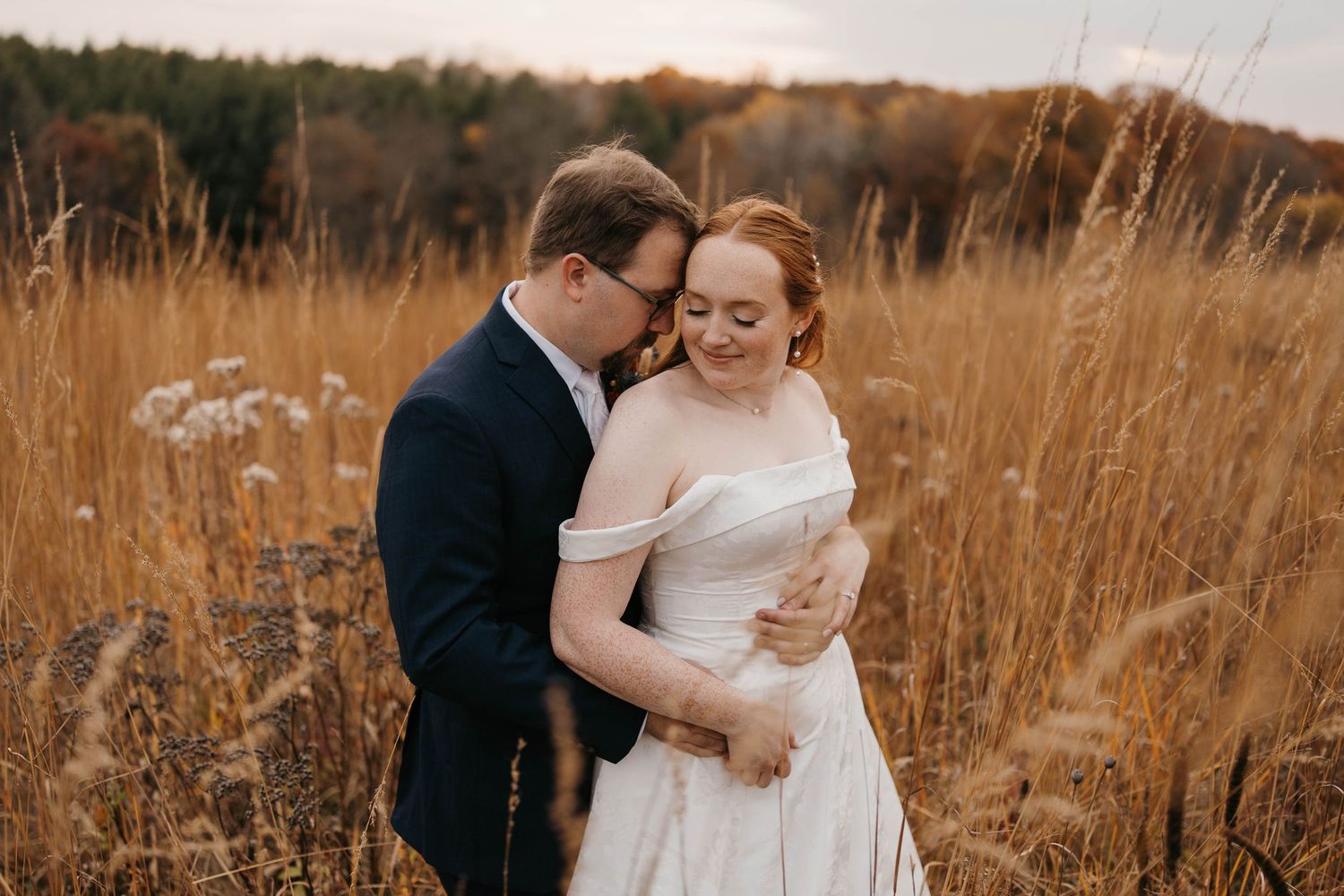 Bride in a white gown embraces a groom in a dark suit while standing in a golden autumn field with tall grass and colorful trees.