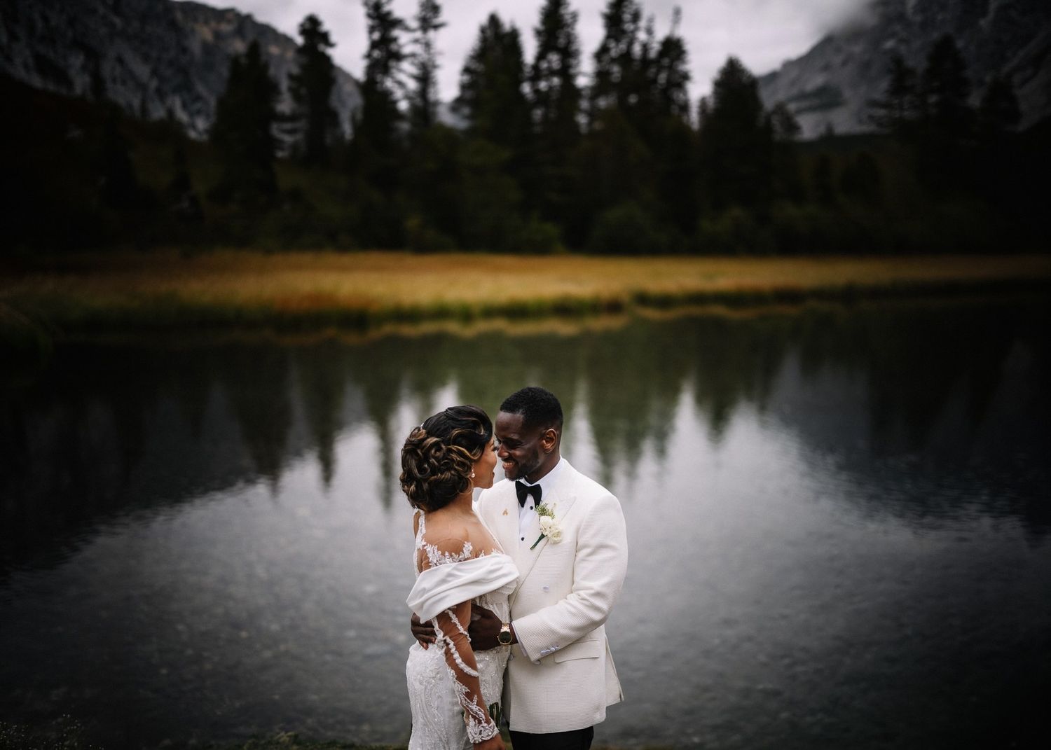 Bride in a white gown and groom in a white tuxedo jacket embrace beside a calm mountain lake with evergreen trees at dusk.